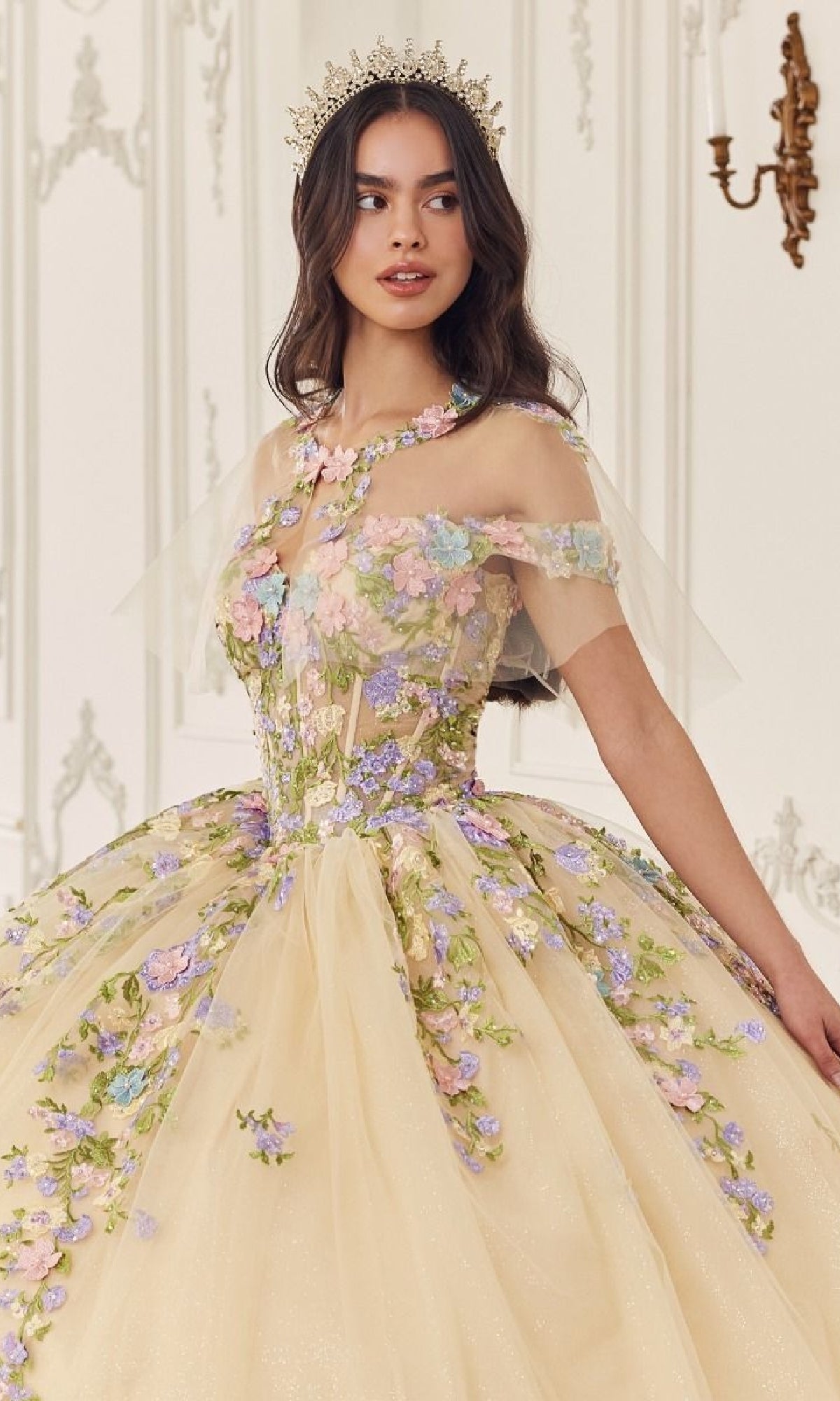 A woman wears the Quinceanera Dress 15724 by Ladivine—a light yellow ball gown with floral embroidery and a sheer overlay—indoors, styled with a decorative crown.
